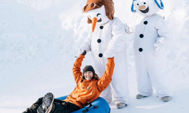 Hymyilevä nainen lumituubissa menossa lumiliukumäkeen. Mukana myös Snowman Worldin lumiukot.